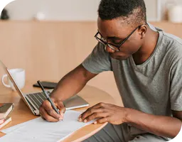 Man writing on paper next to laptop computer